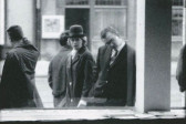 A black and white image of a man looking at documents through a window on a street sidewalk. Behind him, a women in a hat looks at him. People are present walking on the street surrounding them.