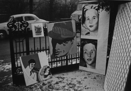 A black and white image of four paintings of various sizes displayed against a fence with a thin layer of snow, marked with footprints, beneath them.