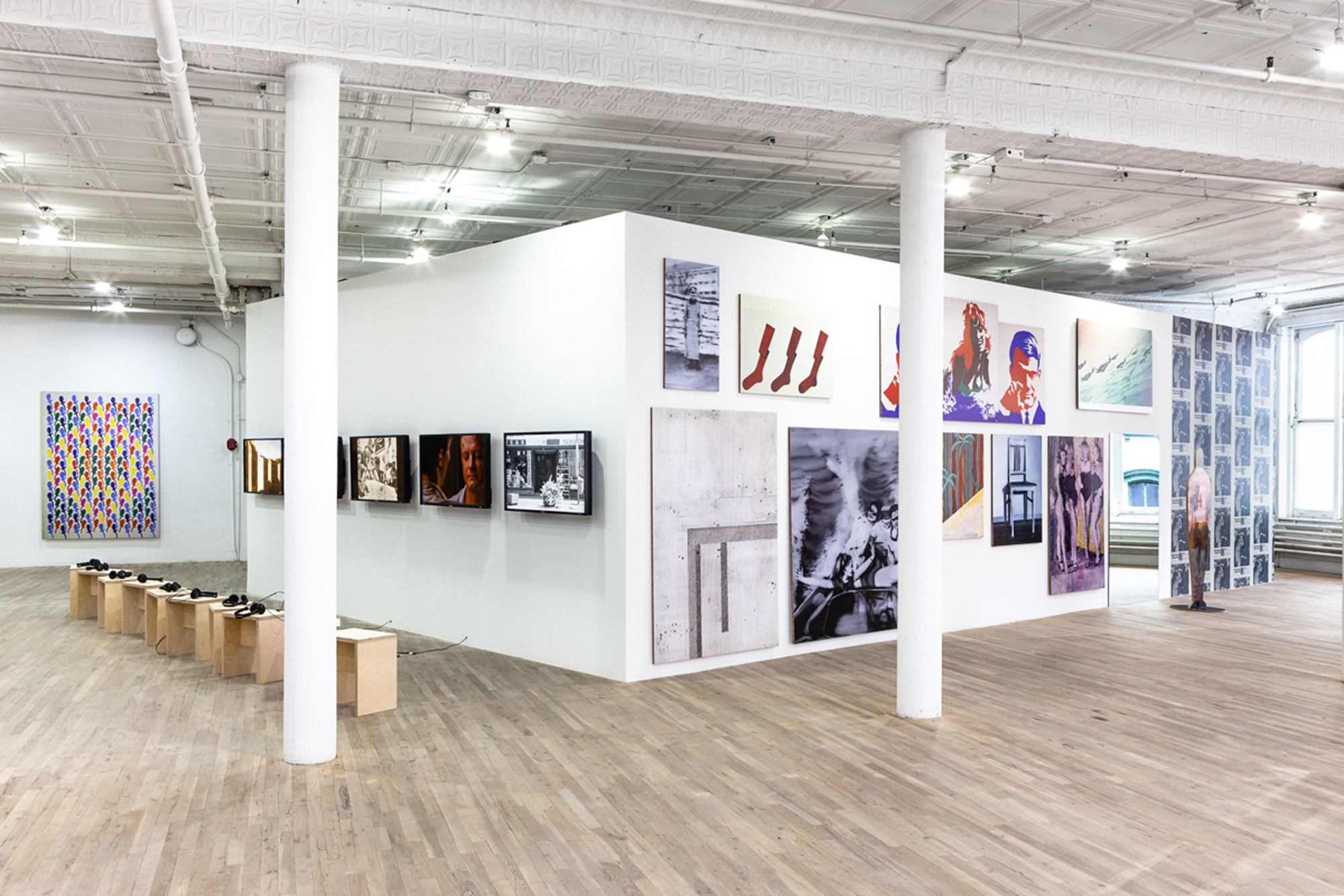 An angled view of the external corner of two walls. On the left are monitors, displaying various images. On the right large paintings and images are hung on all surface area of the wall.