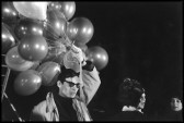 A black and white image of a man in sunglasses holding a large cluster of balloons tied to strings above his head with his left hand.