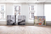 Three large prints rest on the floor against a windowed wall. Two on the left are black and white, of an interior room. On the right is a colorful illustration of two women.