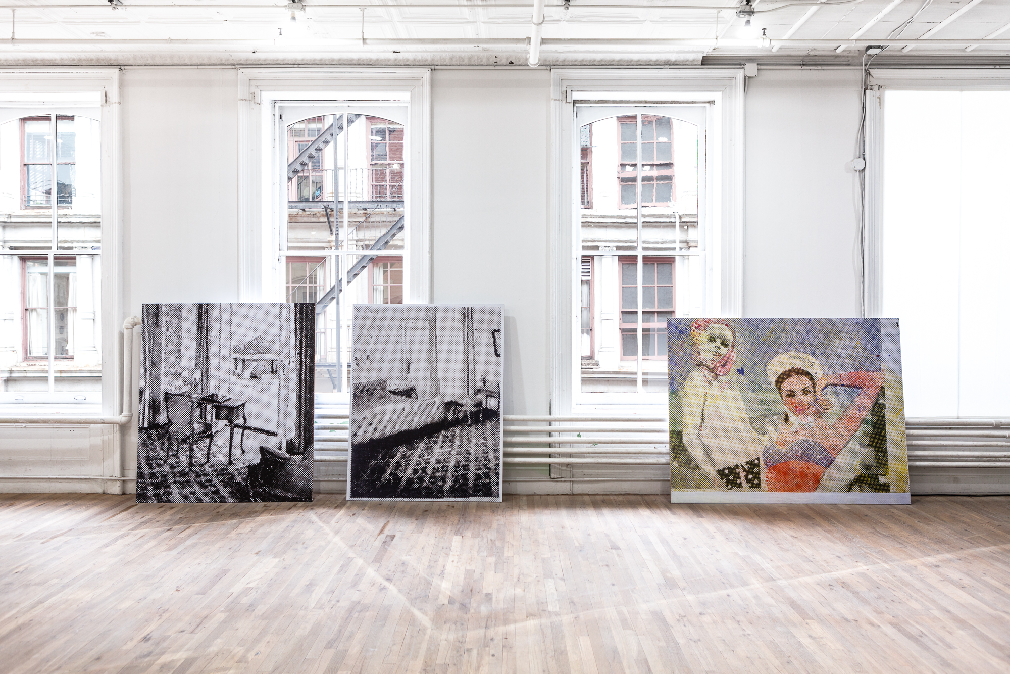 Three large prints rest on the floor against a windowed wall. Two on the left are black and white, of an interior room. On the right is a colorful illustration of two women.