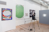 An angled view of a wall portraying four paintings, of various dimensions, at eye level. In the room, a 34mm projector displays a video, to the right, on a gray wall.