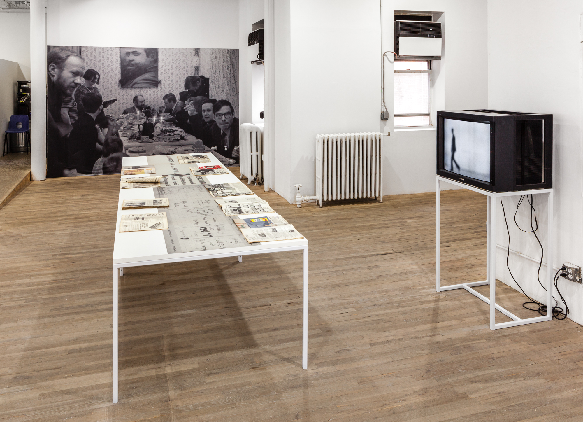 A white table with various documents sits in the ceter for a room. On the right, a televsion displaying a man walking sits on a pedestal. In the background, a black and white image of a crowd at a dinner table is displayed.