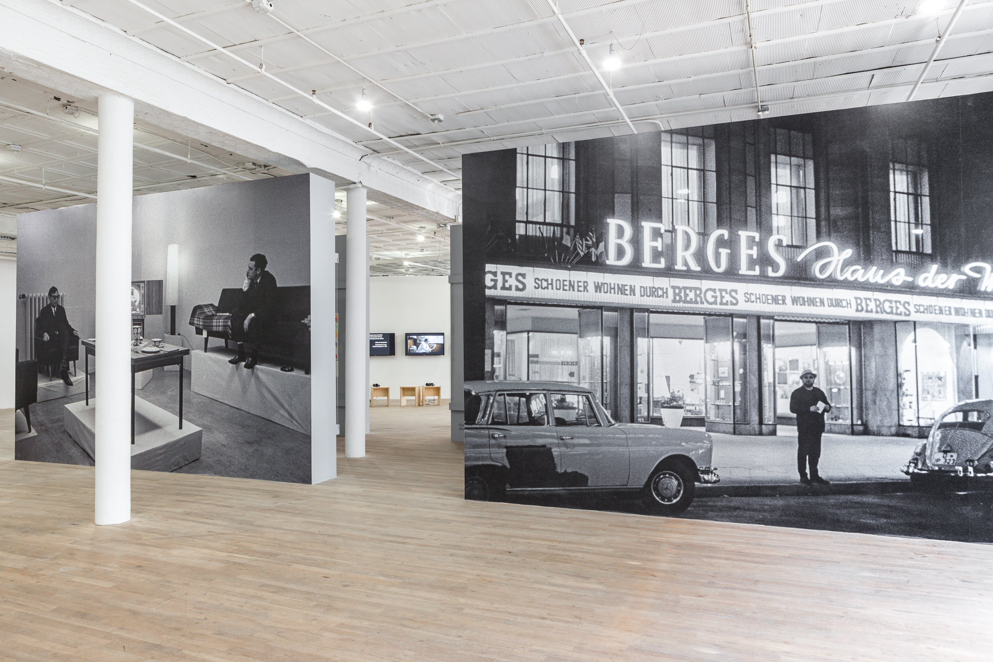 Two walls form an entry way to the exhibition space. Both present black and white photos across their entire surface area. On the left, two men sit in a living room; on the right a man stands outside a store with a neon light sign.