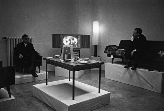 A black and white image of two men sitting in a living room. The furniture is elevated on white pedestals. The man on the left sits on a chair; on the right on a sofa. A table with items sits inbetween them.