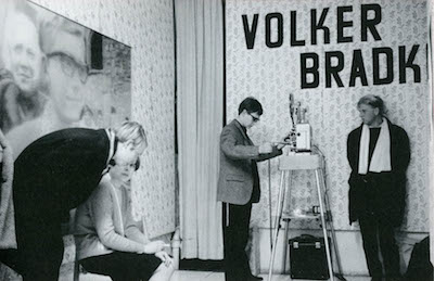 A black and white image of four people, one sitting, three standing in a patterned room displaying a large photograph on the left wall. On the right wall, two men stand underneath large block text, that reads, 
