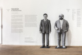 A white wall displaying exhibition text next to two, life size paper mache sculptures. Left is John F. Kennedy and right is gallerist Alfred Schmela. To the right is text thanking the Artists Space suppoters.
