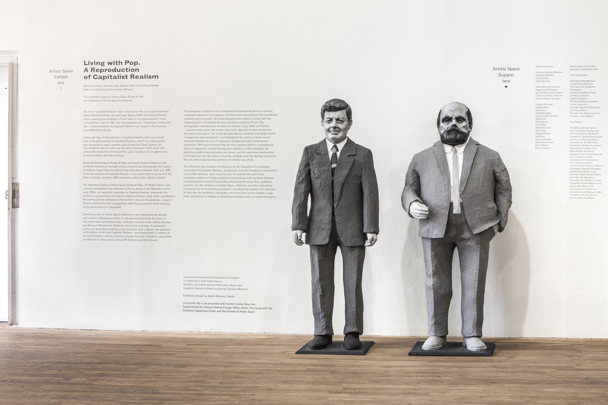 A white wall displaying exhibition text next to two, life size paper mache sculptures. Left is John F. Kennedy and right is gallerist Alfred Schmela. To the right is text thanking the Artists Space suppoters.