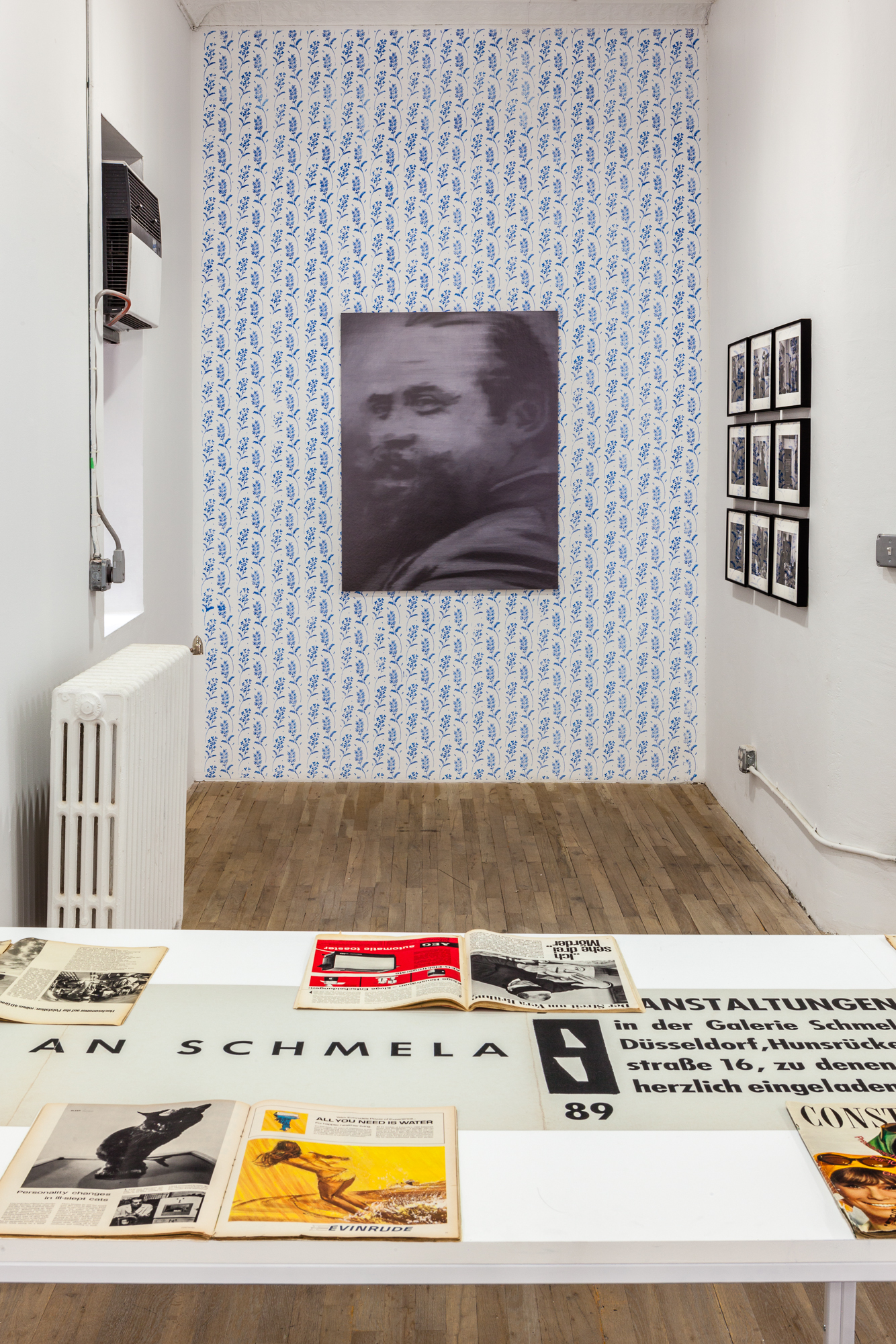 A closed room; in the foreground is a white table with varoius documents. On the back wall hangs a black and white portrait of a man on a blue patterened wall. To the right adjacent wall, hang a grid of photogrpahs.
