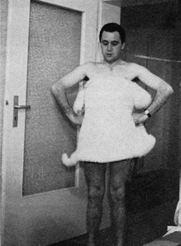 A black and white image of a man, standing in a doorway, coving his torso with a small white blanket.