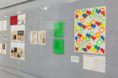A angled view of a grey wall with documents displayed behind glass. The closest image depicts a colorful illustration of shapes, with two, green, square documents to the left of it.