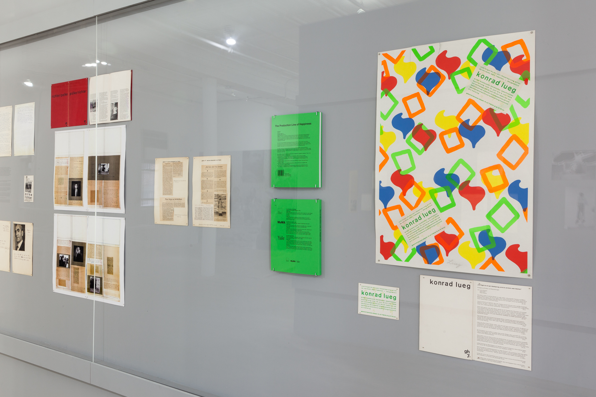 A angled view of a grey wall with documents displayed behind glass. The closest image depicts a colorful illustration of shapes, with two, green, square documents to the left of it.