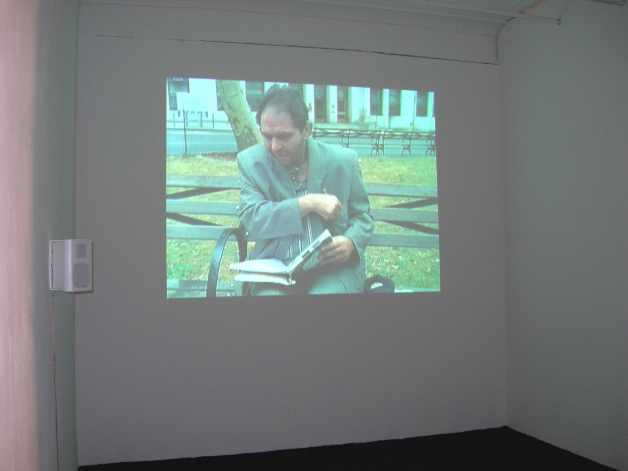 An image of a man sitting on a park bench is projected on a white wall. The man is holding a book open in his lap and is reaching into his breast pocket.
