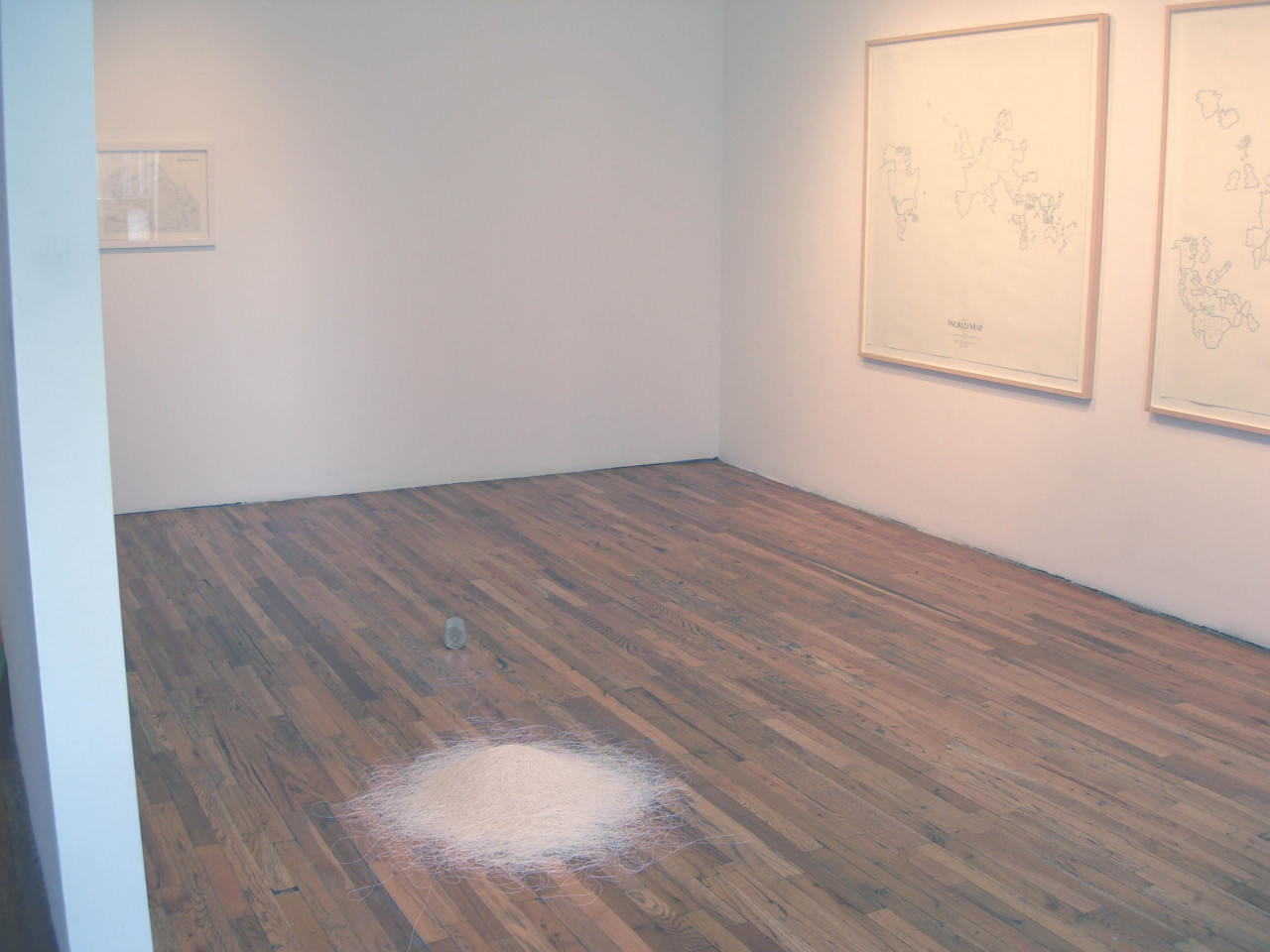 A large pile of string connected at one end to a tin can sits on a wooden gallery floor. On the wall to the right are two framed world maps in black and white.