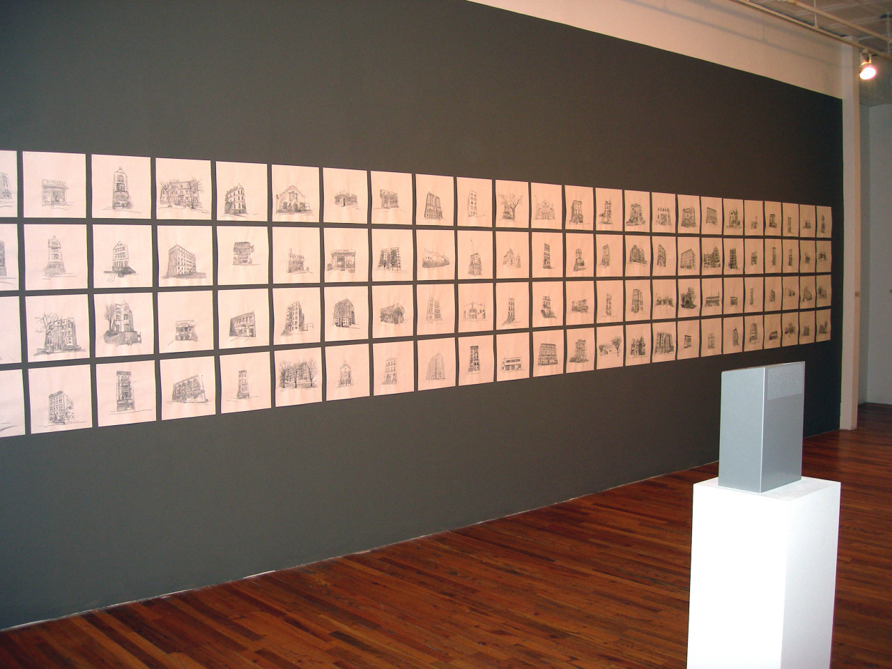 A gray wall with dozens of drawings arranged in rows of four. The drawings depict single buildings in black on white paper. In front of the wall, a gray object stands on a white plinth.