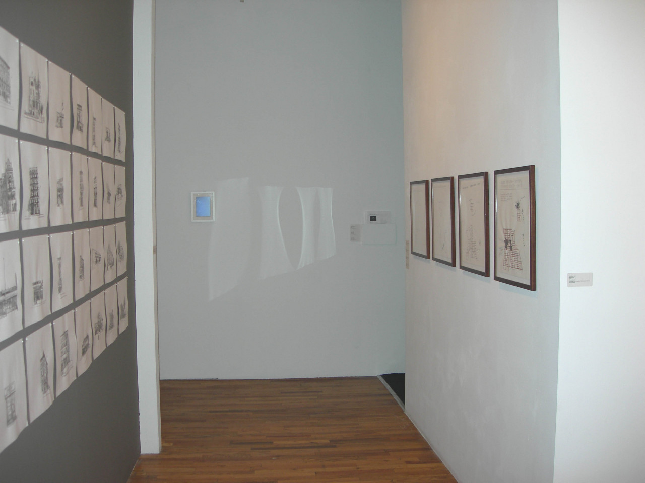 A hallway with one gray wall and one white. On the gray wall, there are a series of drawings on white paper arranged in a 4x7 grid. On the white wall, there are four frames. Light is refracted on a third wall at the end of the hallway.