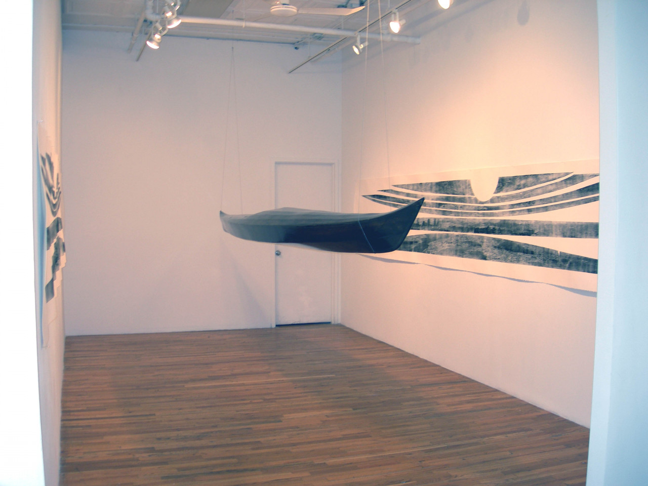 A black, kayak-shaped sculpture is suspended from the ceiling in a gallery space. On either wall, there are black and white prints mimicking the structure of the boat.