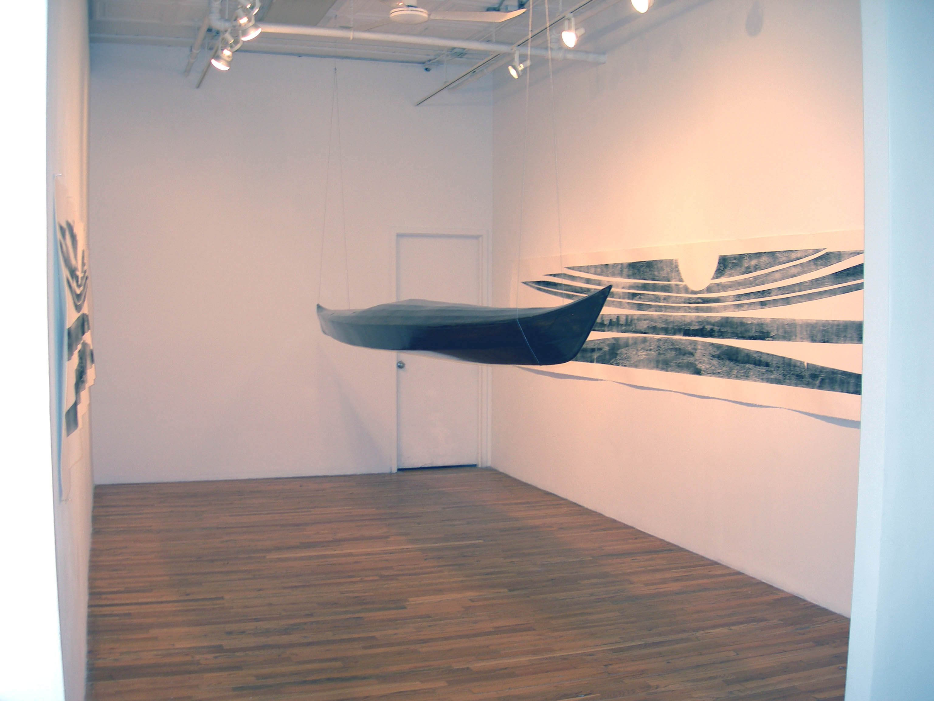 A black, kayak-shaped sculpture is suspended from the ceiling in a gallery space. On either wall, there are black and white prints mimicking the structure of the boat.