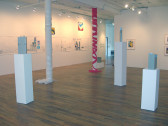 A gallery space with two columns. One column is wrapped in an off-white fabric, the other in bright pink. There are four white plinths of various heights with gray objects on top. One of the surrounding walls has a long sheet of paper with a drawing of highway signs and a gas station on it; the other has colorful framed images.