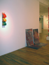 Three red, orange, and green fabric rolls are arranged vertically on a white wall. Further along the wall, two marbled, gray and red sheets have been hung low on the wall such that their lower halves drape on the gallery floor.