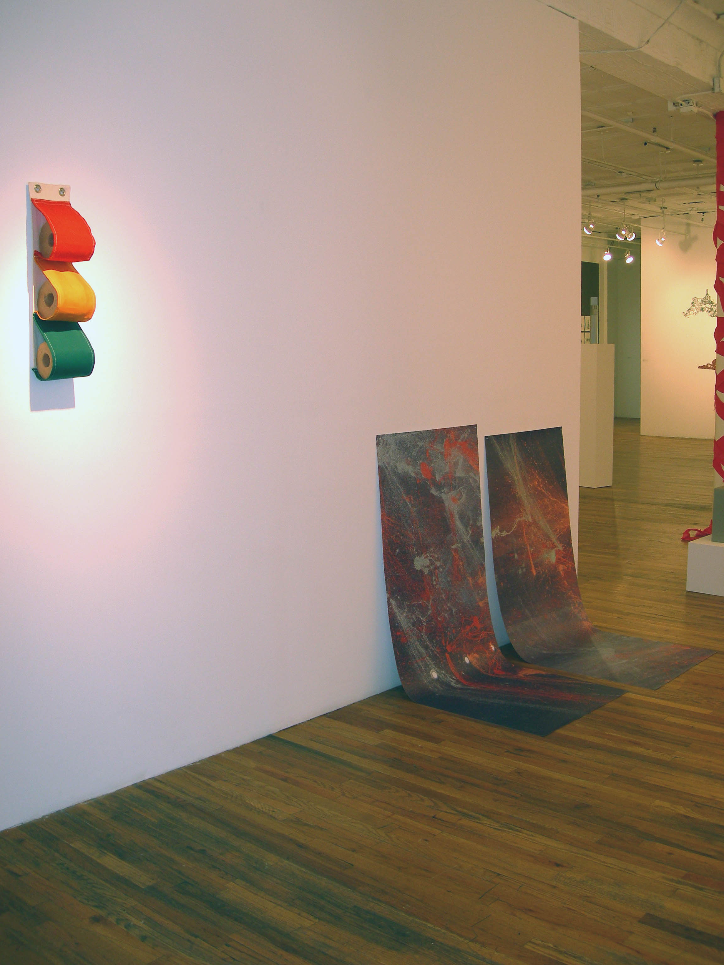 Three red, orange, and green fabric rolls are arranged vertically on a white wall. Further along the wall, two marbled, gray and red sheets have been hung low on the wall such that their lower halves drape on the gallery floor.
