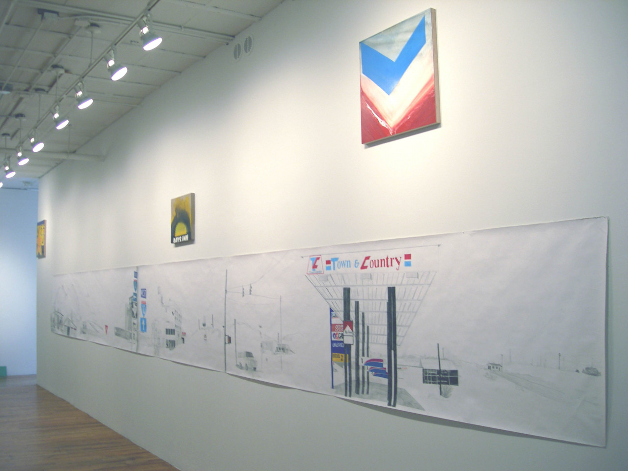 A long sheet of paper with a drawing of highway signs and a gas station on it hanging on a white wall. Hung above that are three paintings of logos.