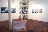 Several sculptures, paintings, video works, and photographs installed in a gallery space with wooden flooring. A sculpture consisting of a white panel affixed to a wooden board upon which two striped rocks rest is installed on the ground. A TV monitor is installed in the room's corner directly behind the sculpture. The portion of the wall directly behind the TV monitor has been covered with a grid of poster-like images. Three color photographs and two paintings with muted purple and gray tones frame the monitor on its left and right sides.