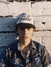 Photographic portrait of a youthful figure leaning against a white brick wall. They wear a partially unbuttoned, blue and black plaid shirt and a trucker hat. Large black text emblazoned across the front of the hat reads, 