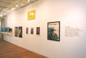Several photographs and TV monitors installed along the length of a white wall in a gallery space. Black vinyl text on the wall's far right side reads, 