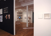 Several sculptures and 2D works installed in a gallery space with wooden flooring. Two walls emerge on the left and right sides of the image, creating a doorway through which another area of the gallery is visible. The wall on the left is painted black, while all other walls are painted white. Several photographs—including two picturing nude figures bound in rope—and works on paper hang on either wall. In the far gallery space, three sculptures sit on the floor and a single TV monitor is installed on the farthest wall.