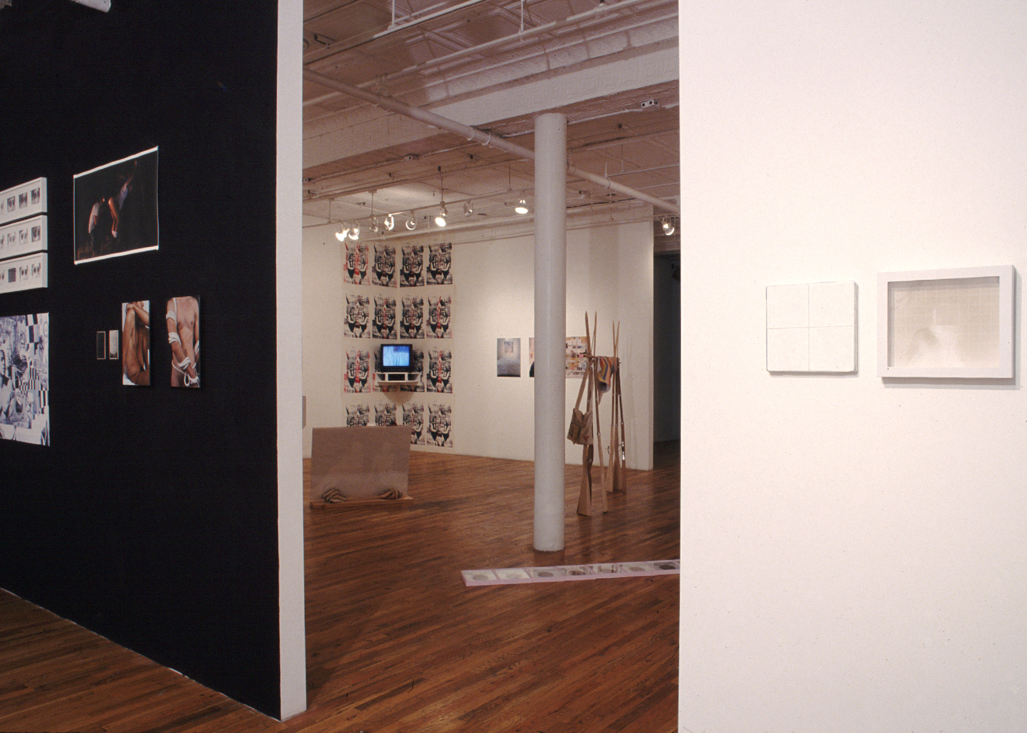 Several sculptures and 2D works installed in a gallery space with wooden flooring. Two walls emerge on the left and right sides of the image, creating a doorway through which another area of the gallery is visible. The wall on the left is painted black, while all other walls are painted white. Several photographs—including two picturing nude figures bound in rope—and works on paper hang on either wall. In the far gallery space, three sculptures sit on the floor and a single TV monitor is installed on the farthest wall.
