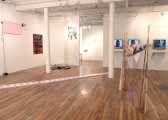 Several sculptures, video works, and other artworks installed in a gallery space with wooden flooring. A large sculpture on the image's right side is composed of a series of wooden rifles gathered to form two, pole-like structures, between which a quilt-like blanket is wrapped around a supporting pole. To the left of this sculpture, a second, long structure composed of a series of white tiles runs diagonally across the gallery floor, forming a long line. Several TV monitors and 2D works are installed on the gallery walls behind the two sculptures.