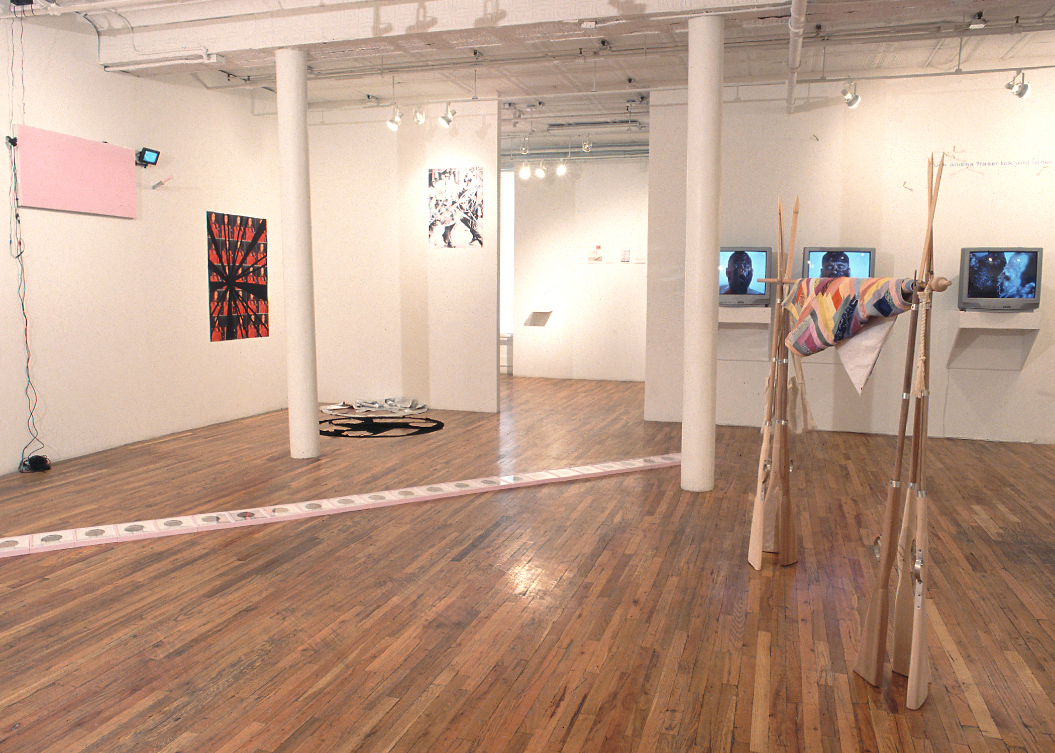 Several sculptures, video works, and other artworks installed in a gallery space with wooden flooring. A large sculpture on the image's right side is composed of a series of wooden rifles gathered to form two, pole-like structures, between which a quilt-like blanket is wrapped around a supporting pole. To the left of this sculpture, a second, long structure composed of a series of white tiles runs diagonally across the gallery floor, forming a long line. Several TV monitors and 2D works are installed on the gallery walls behind the two sculptures.