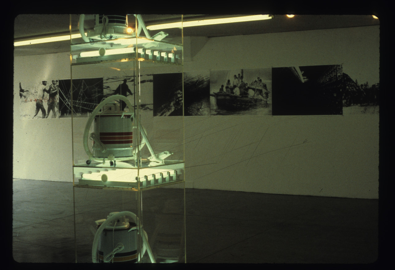 In the foreground vacuums on plexiglass shelves with fluorescent lights underneath are visible and in the background on a white gallery wall is a number of different photographs lined next to each other in successio.