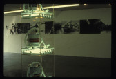 In the foreground vacuums on plexiglass shelves with fluorescent lights underneath are visible and in the background on a white gallery wall is a number of different photographs lined next to each other in successio.