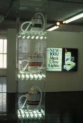 In the foreground is a set of vacuum cleaners on display vertically above each other on a plexiglass shelf rack with fluourescent lights underneath each one. Behind it on a wall is a Duratrans on a lightbox with text that reads: 