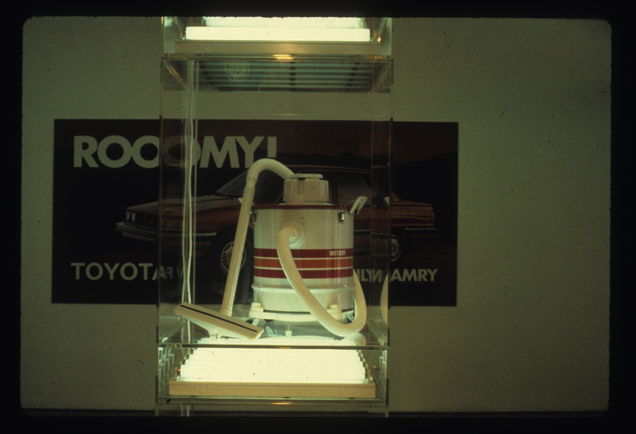 In the foreground a vacuum can be seen on a plexiglass shelf case with a fluorescent light underneath and above it. Behind it is an image on a white wall of an automobile with text visible that reads, "ROOOMY!" on the top left and "TOYOTA" on the bottom left and "NEW FAMILY CAMRY" on the bottom right.