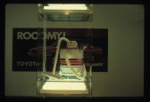 In the foreground a vacuum can be seen on a plexiglass shelf case with a fluorescent light underneath and above it. Behind it is an image on a white wall of an automobile with text visible that reads, 