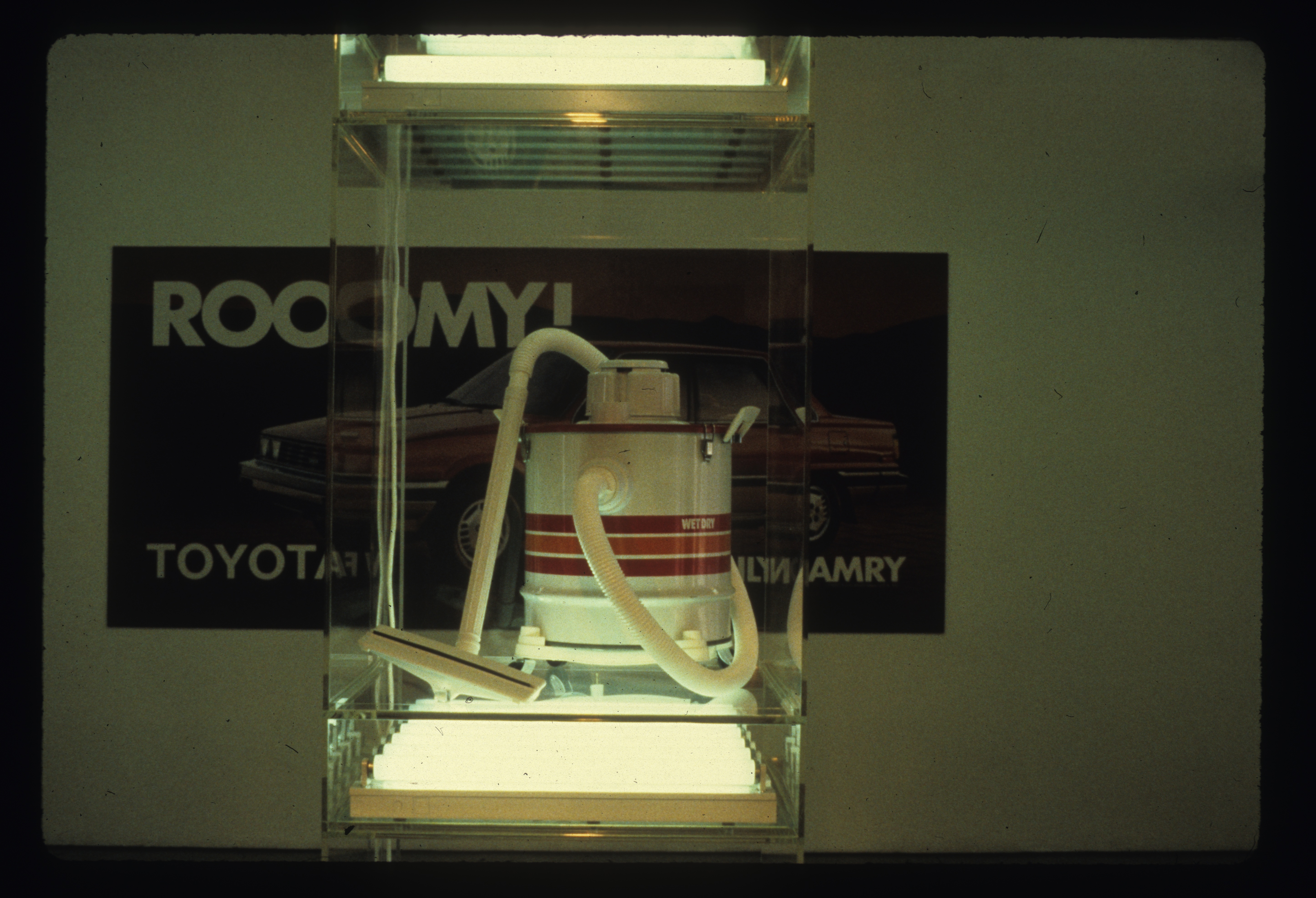 In the foreground a vacuum can be seen on a plexiglass shelf case with a fluorescent light underneath and above it. Behind it is an image on a white wall of an automobile with text visible that reads, 