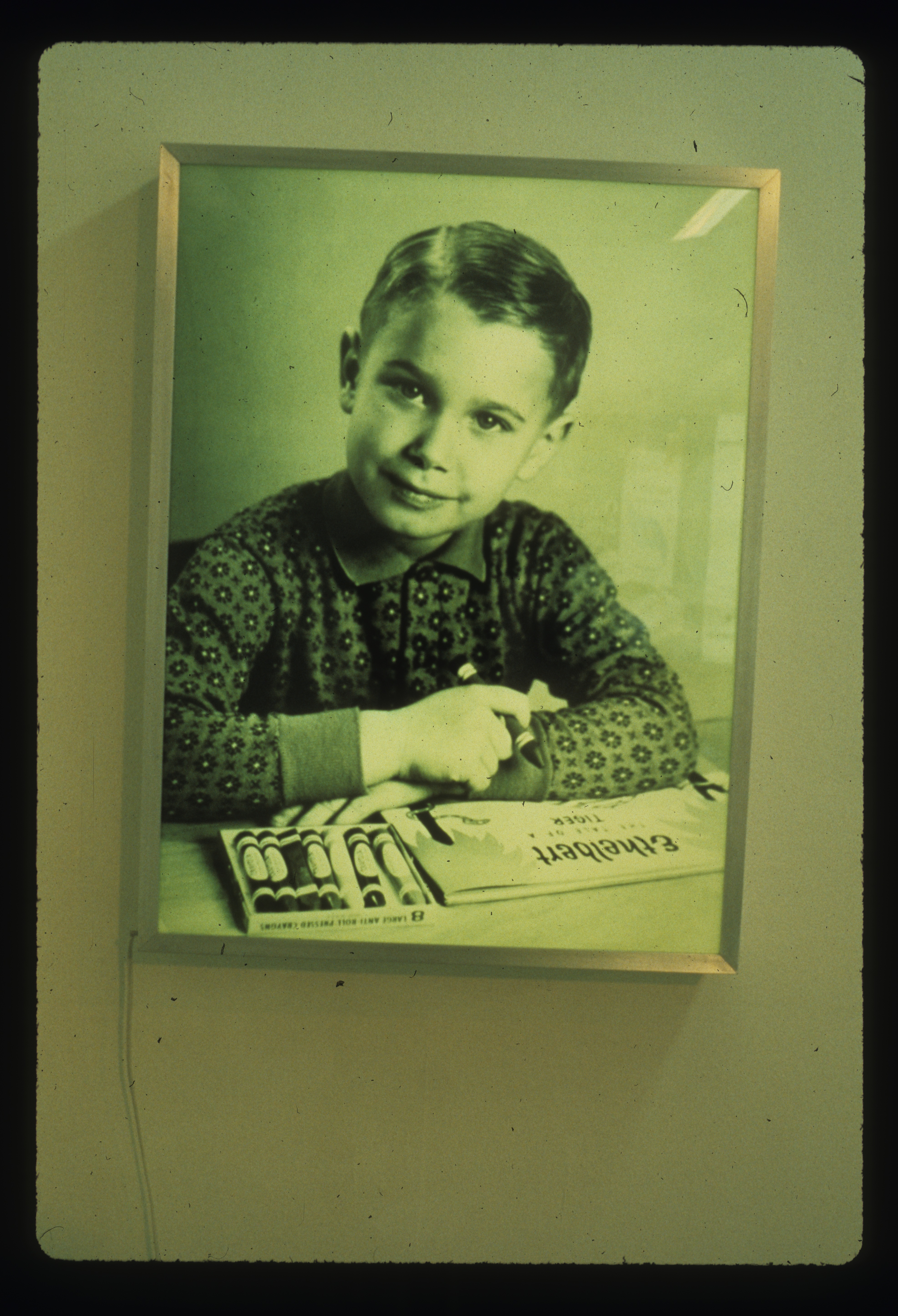 A black and white Duratrans on a lightbox of a portrait of a child holding a crayon with a box of crayons and a small book lie on a desk in front of them under their crossed arms. This is on a wall with the plug for the box coming from under the left side.