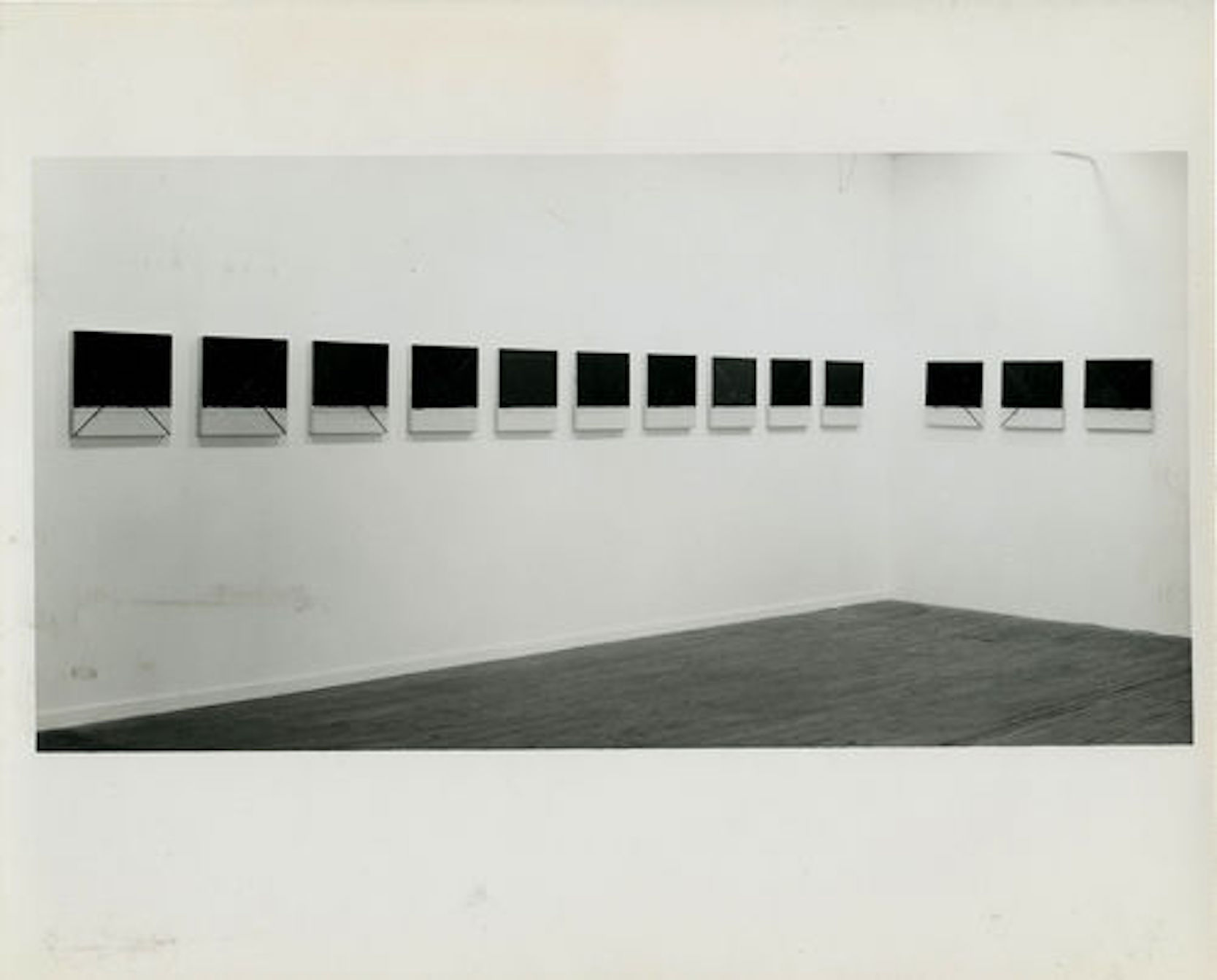 A black and white installation photograph of thirteen dark squares hung in a gallery space.