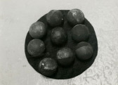 A black and white photograph of a sculpture comprised of nine dark orbs on a dark circular backgroun.