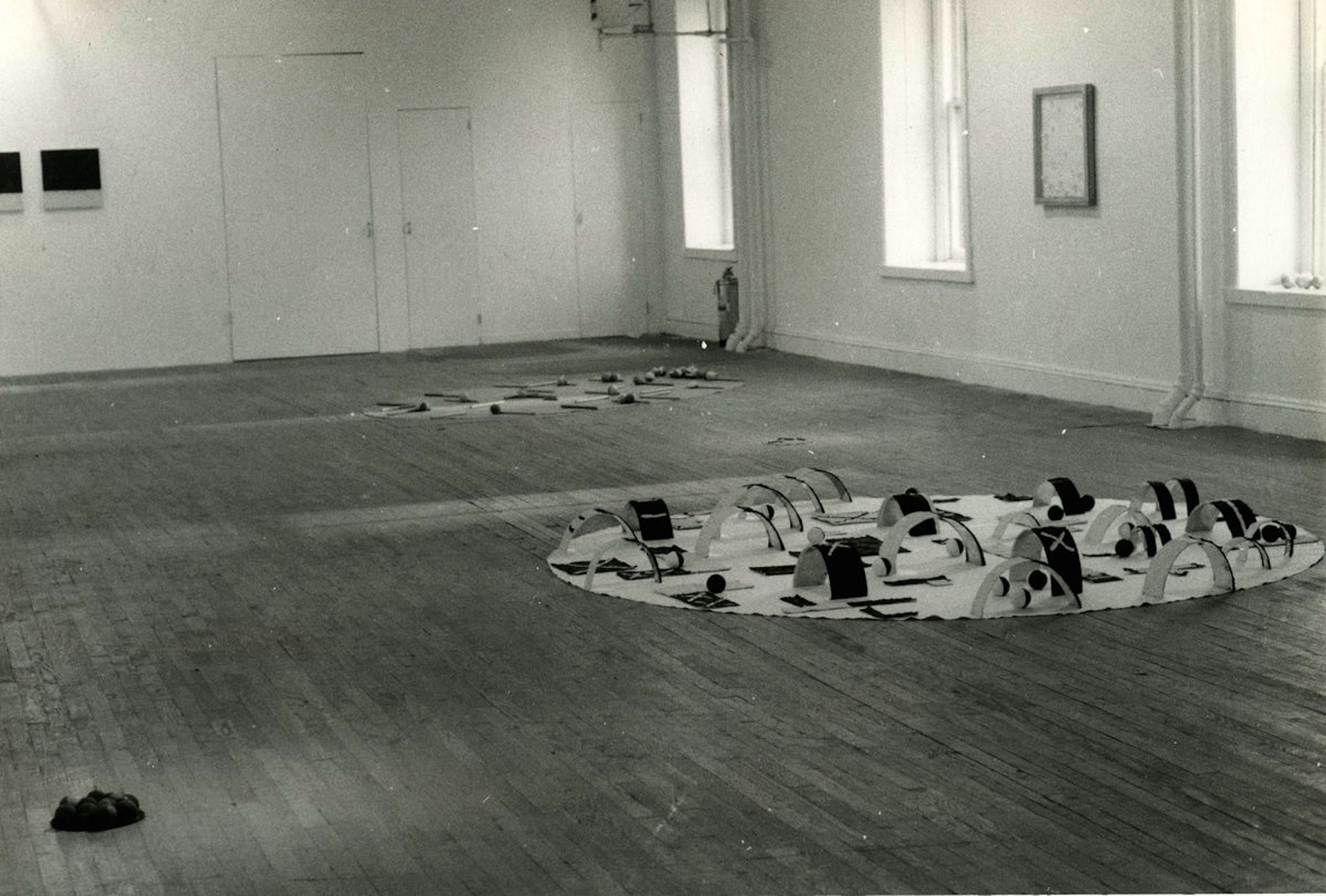 A black and white installation photograph of a gallery space. Two floor bound sculptures are visible in the foreground, and in the background a few framed works hangfrom the wall.