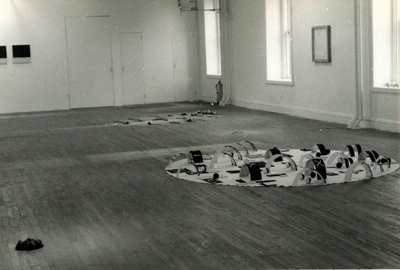A black and white installation photograph of a gallery space. Two floor bound sculptures are visible in the foreground, and in the background a few framed works hangfrom the wall.