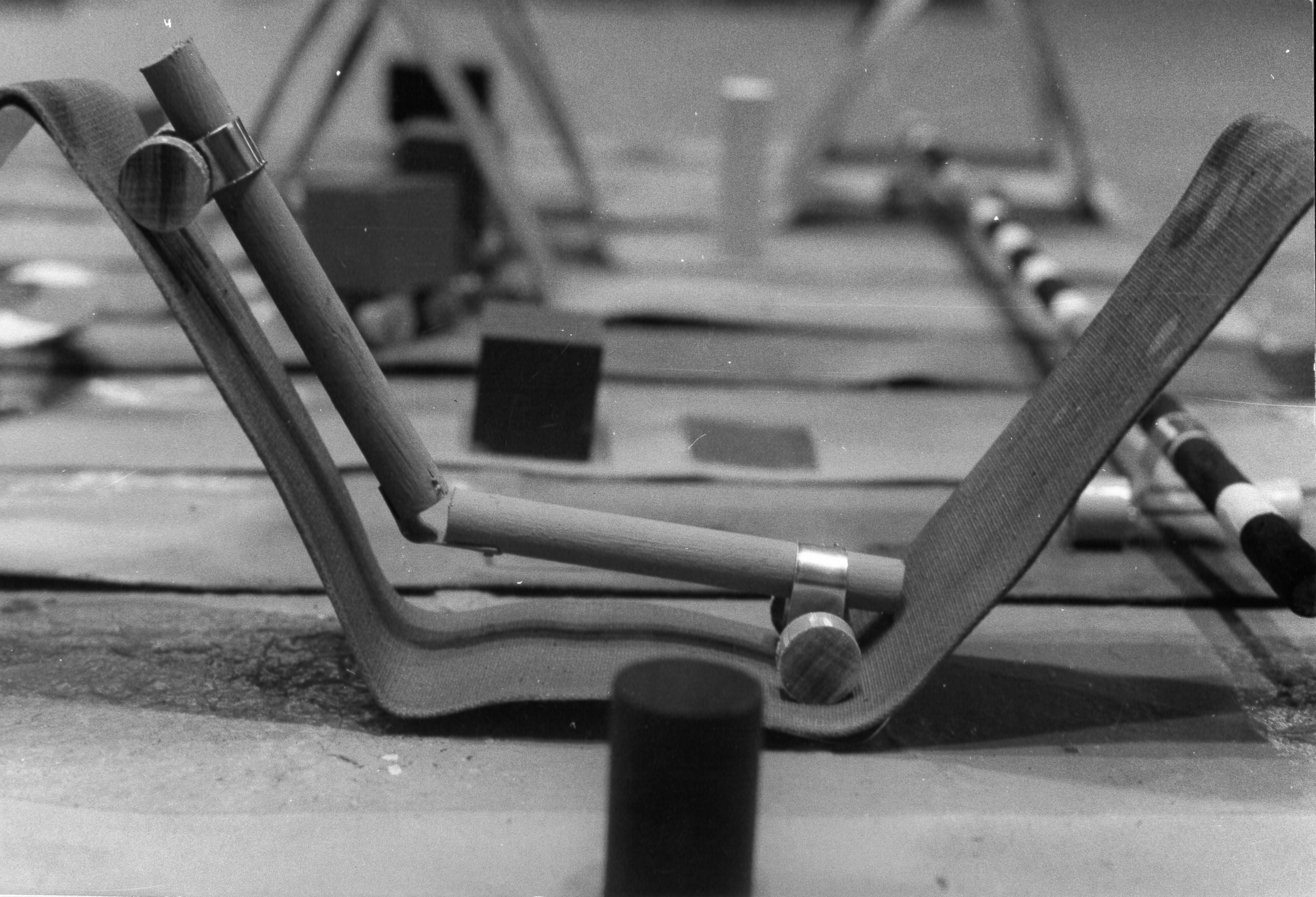 A black and white installation photograph of a sculpture. The work features two wooden rods, connected to form an obtuse angle in the center. The end of each rod has a set of wheels, and the piece rests on a sling-like piece of sturdy fabric.