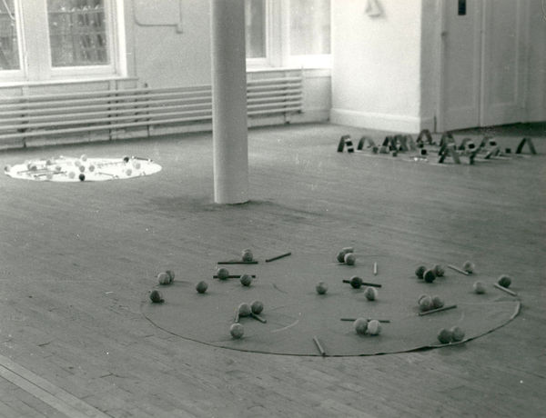 A black and white installation photograph of a gallery space. Placed in a triangle formation around a column, several small objects lie on circular or rectangular mats on the floor.