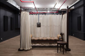 An off-white curtain hangs from a wooden four-poster bed frame. One side of the curtain is partially open, revealing a patterned chaise lounge and a framed image on a blue chain.