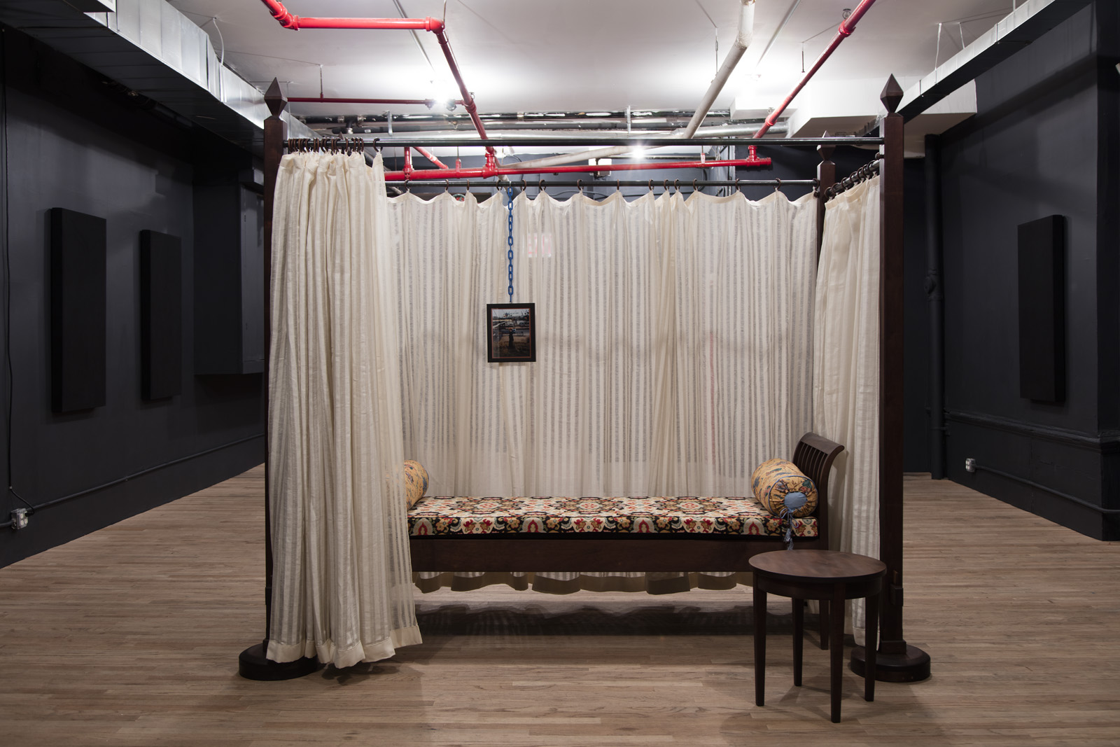 An off-white curtain hangs from a wooden four-poster bed frame. One side of the curtain is partially open, revealing a patterned chaise lounge and a framed image on a blue chain.