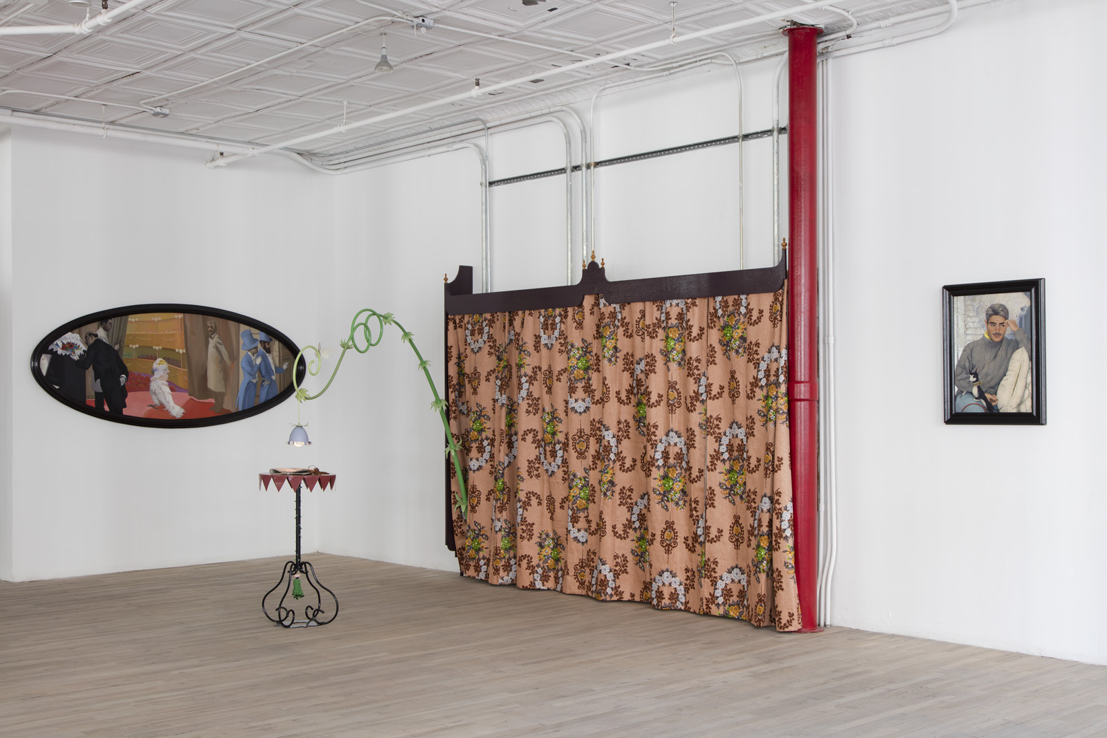 Two paintings and several sculptural elements are installed in a corner. The paintings, one in an oval frame and the other rectangular, have figurative subjects of humans and animals. A whimsical end table is illuminated by a light fixture shaped like a flowering plant whose stem extends from a patterned curtain hung against the wall.